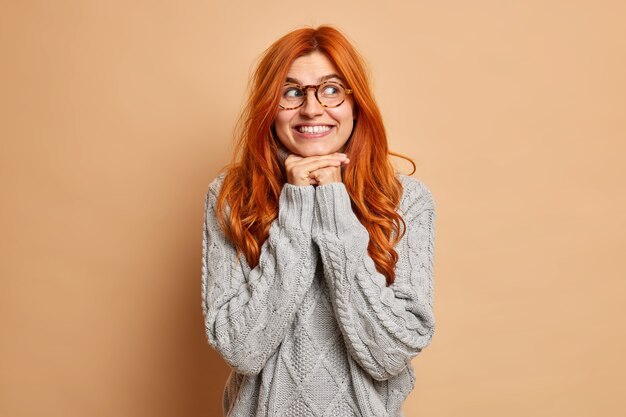 Good looking young woman with red hair keeps hands under chin smiles toothily concentrated aside wears spectacles and sweater.