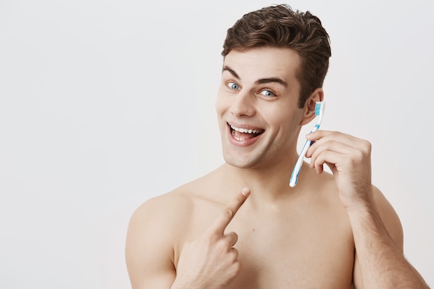 Good-loooking muscular guy with healthy skin, woke up early, smiling broadly, having fun indoors, holding toothbrush pretending to use it as a cell phone, pointing with index finger at toothbrush.