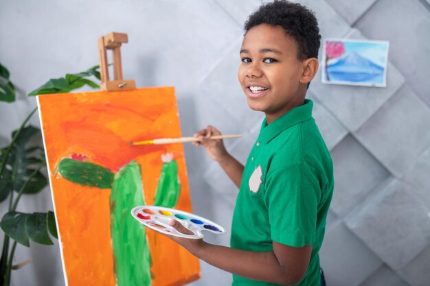 Good moments. Happy dark-skinned school age boy holding palette, painting cactus on easel, looking head turned at camera in light room