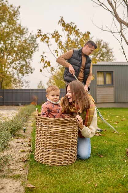 Good moments. Profile of young joyful pretty woman in warm clothes crouching looking at child in basket smiling at camera and man raking leaves in garden