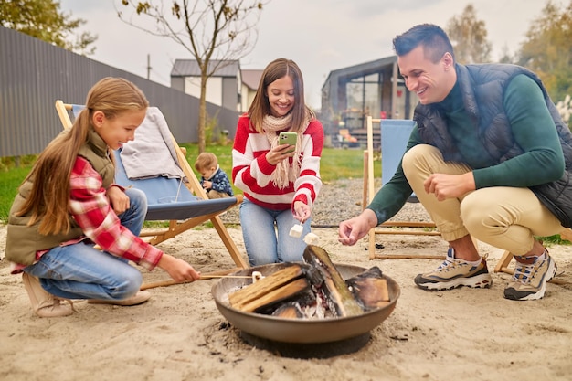 Free photo good moments. smiling young adult man in casual clothes pretty woman with smartphone and school age girl crouching holding marshmallows on stick over campfire in courtyard