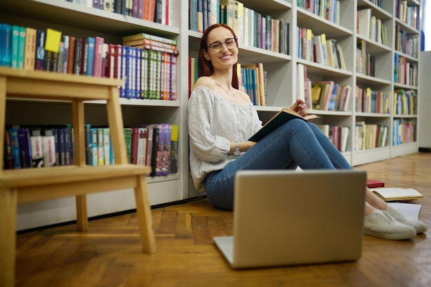 Good moments. Smiling young pretty woman in glasses with book sitting on floor near laptop leaning on bookshelves looking at camera