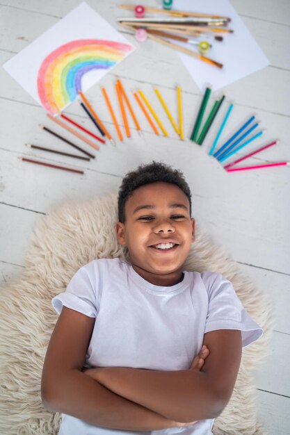 Good moments. Top view of dark-skinned smiling school-age boy in white tshirt lying on floor with folded hands and near colored pencils, brushes and drawing in bright room