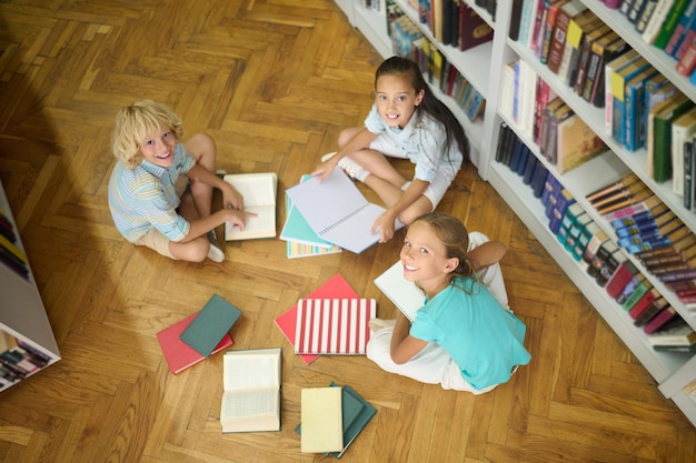 Good moments. Top view of smiling boy and two girls of school age sitting on floor of library holding faces up and looking at camera