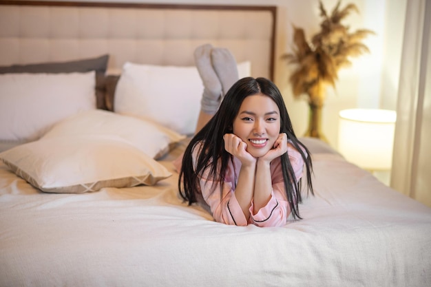 Хорошее настроение. Довольно улыбающаяся молодая азиатка с длинными темными волосами в розовой пижаме лежит на кровати, касаясь руками подбородка, глядя в камеру дома в спальне