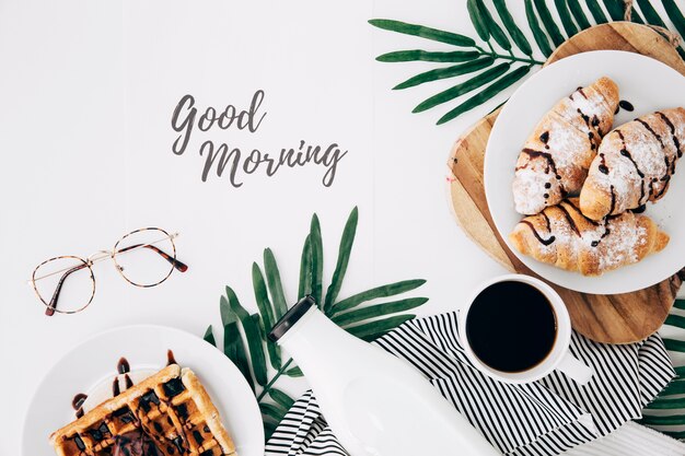 Good morning text with eyeglasses; fresh baked croissant; waffles; bottle and coffee cup on white desk