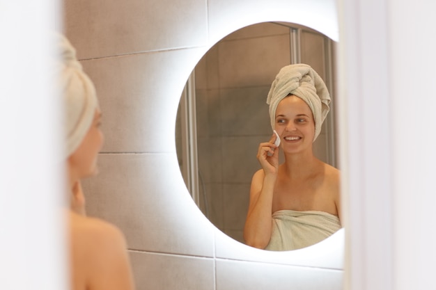 Una buona giovane donna adulta avvolta in un asciugamano bianco in posa in bagno davanti allo specchio pulisce il viso con un batuffolo di cotone, sembra felice e sorridente, cura della pelle, routine mattutina.