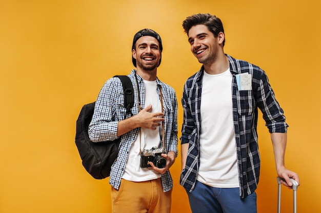 Добродушные молодые брюнеты в клетчатых рубашках и белых футболках улыбаются на оранжевом фоне Веселые путешественники позируют с ретро-камерой и рюкзаком