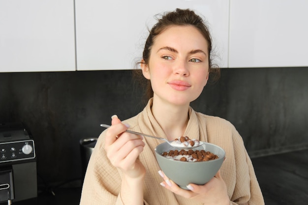 Бесплатное фото Красивая брюнетка ест завтрак, стоя на кухне возле столешницы и держа в руках миску