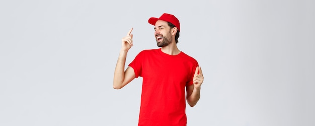 Goodlooking carefree bearded man in red tshirt and cap uniform of employee close eyes and dancing po...