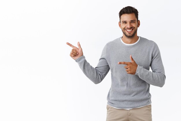 Goodlooking charming bearded caucasian guy with short stylish hairstyle wear grey sweater pointing left advertise product corporate promotion give advice or recommendation smiling pleased