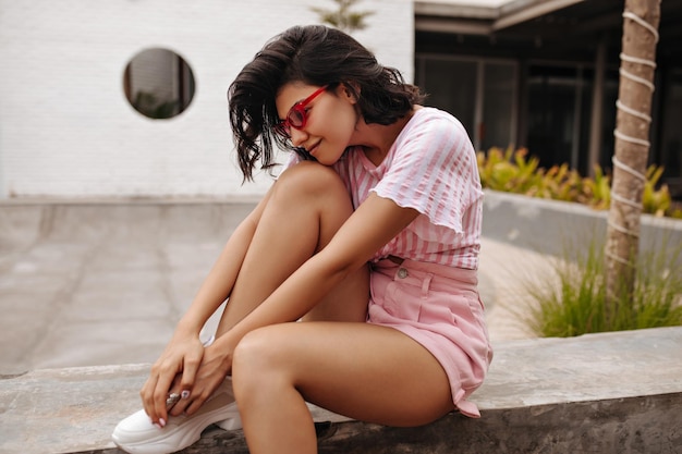 Free photo goodlooking girl in shorts sitting on urban background outdoor shot of cute young woman with tanned skin wearing pink sunglasses