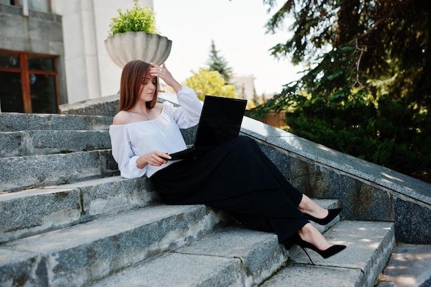 Красивая молодая женщина в белой блузке, широких черных брюках и черных классических высоких каблуках сидит на лестнице и работает на своем ноутбуке
