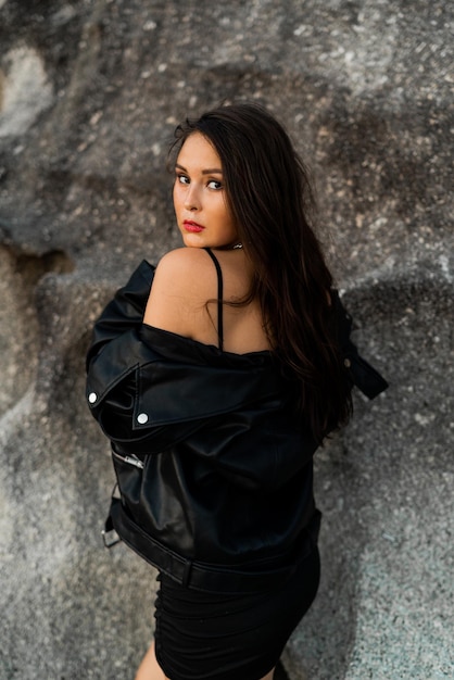 Gorgeous asian woman with red lips in trendy black leather jacket and sensual black dress posing on rocky background