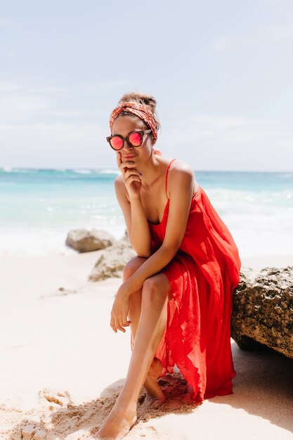 Gorgeous barefooted girl sitting on stone with pensive face expression. Outdoor shot of charming tanned lady in red posing with pleasure on sea.