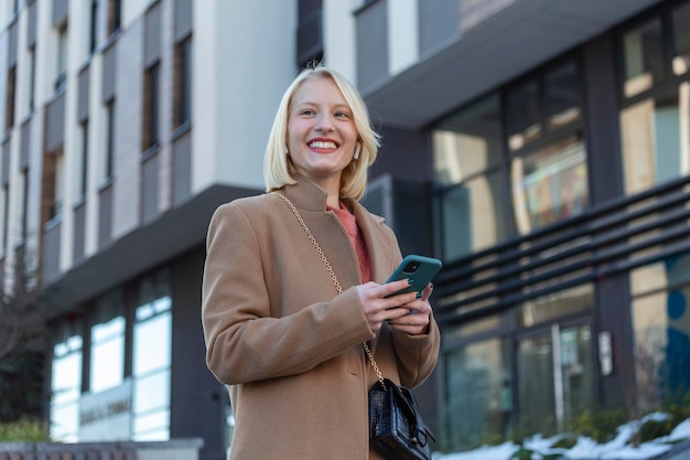 Великолепная красивая молодая женщина со светлыми волосами обменивается сообщениями на смартфоне на фоне городской улицы