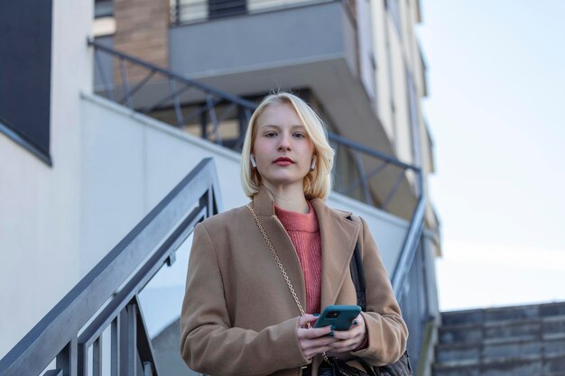 街の通りの背景でスマートフォンでブロンドの髪のメッセージとスマートフォンの会話をしているかわいい女の子とゴージャスな美しい若い女性