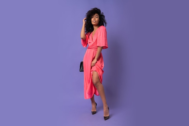 Gorgeous black woman in pink  party dress posing   over  purple wall.  Wearing heels. Full lenght.