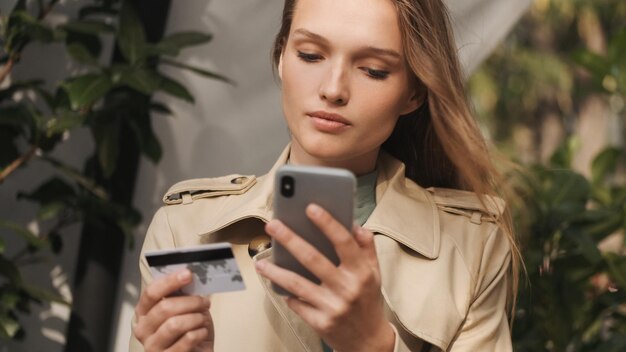 Gorgeous blond girl looking concentrated paying by credit card using smartphone for it during coffee break in street cafe Online purchases concept