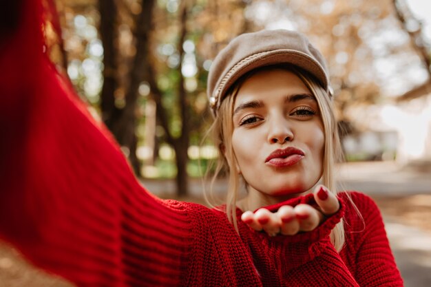 화려한 금발 소녀는 공원에서 그녀의 셀카에 키스를합니다. 사랑스러운 여자는 세련된 빨간 스웨터와 가벼운 모자를 착용합니다.