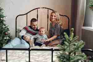 Free photo gorgeous blonde lady with her loving husband and cute kid sitting on a bed near christmas tree