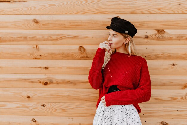 검은 모자와 나무 벽에 다 정하게 포즈 좋은 빨간 스웨터에 화려한 금발 여자. 밖에 서 따뜻한가 날을 즐기는 매력적인 소녀.