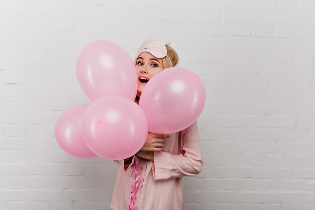Gorgeous blonde woman in pink pajama holding balloons on white wall. Indoor portrait of stunning relaxing birthday girl enjoying celebration.