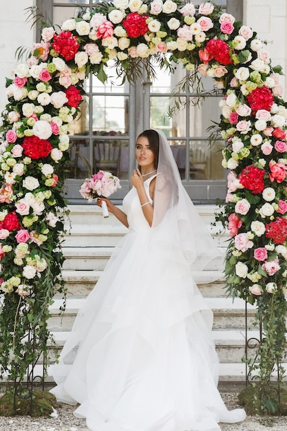 Gorgeous bride stands under wedding altar made of red and white flowers