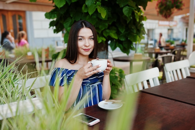 Великолепная брюнетка сидит на столе в кафе с чашкой кофе и напитком