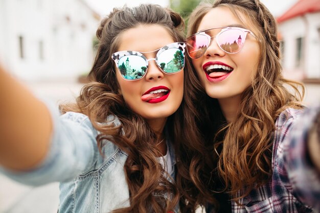 Gorgeous brunette girlfriends with hairstyle, mirrored sunglasses and red lips making selfie with duck face.
