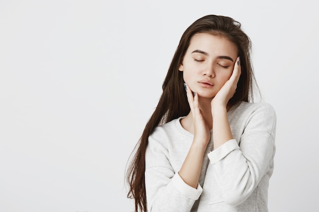 Gorgeous brunette woman with long loose hairstyle wearing long-sleeved t-shirt with closed eyes, keeping hands ob cheeks. Beautiful tender young female of European appearance posing indoors