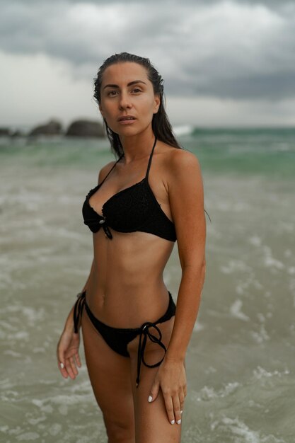 세련된 검은 수영복을 입고 열대 해변에서 포즈를 취하는 완벽한 그림을 가진 화려한 갈색 머리 여자