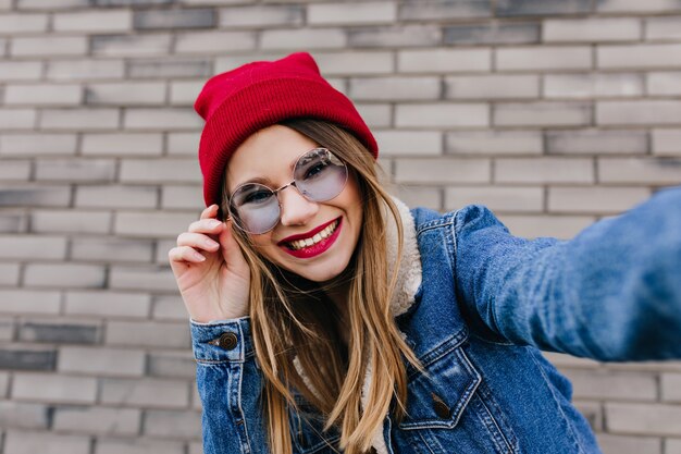 Gorgeous caucasian female model playfully touching her sunglasses. Outdoor shot of laughing fashionable woman with light hair posing near brick wall.