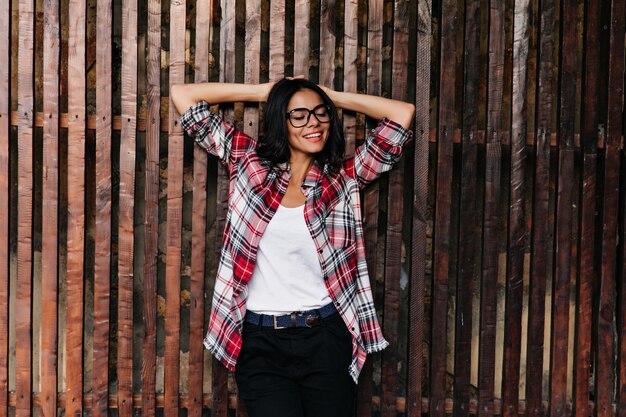 Gorgeous caucasian lady in casual spring outfit enjoying good day. Outdoor photo of relaxed young woman in glasses standing on wooden wall.