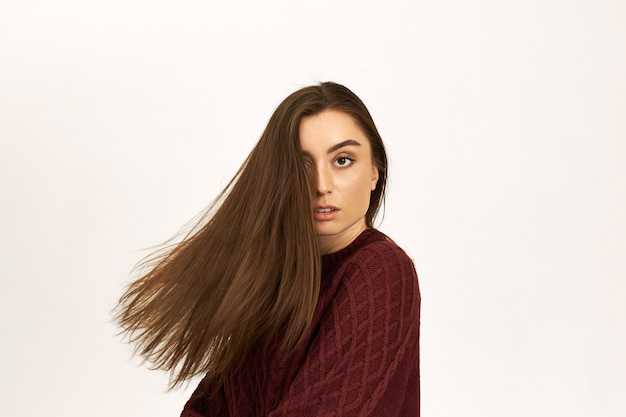 Gorgeous confident young female in warm jumper posing isolated advertising shampoo, turning head, her beautiful shiny hair flying away.
