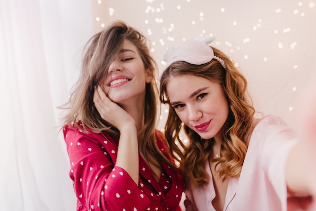 Gorgeous curly woman in sleepmask making selfie in morning with sister. Amazing brunette girl in red pajamas smiling while her friend taking picture.