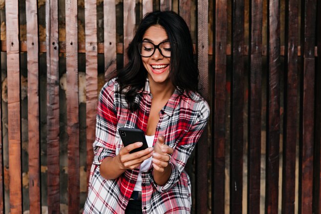 Gorgeous dark-haired woman looking at phone screen and laughing. Outdoor portrait of beautiful latin girl isolated on wooden wall with her device.