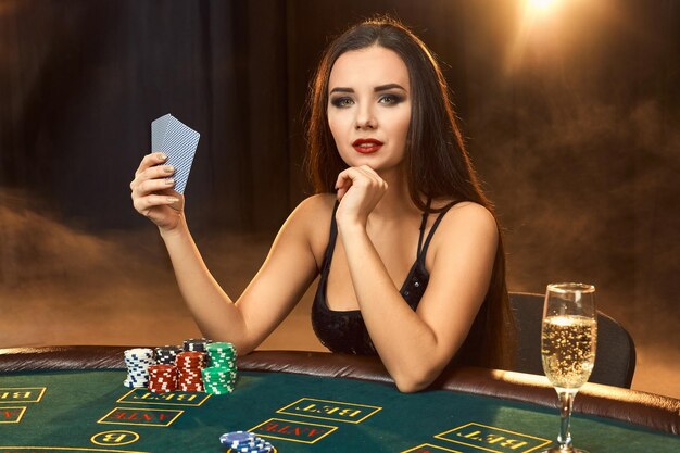 シャンパングラスとポーカーテーブルに座っている黒いドレスを着たゴージャスな感情的な若い女性。煙をしましょう。ポーカー。カジノ