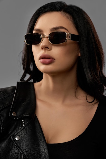 Free photo gorgeous fashion model in trendy sunglasses, bodywear and leather jacket posing over grey background. caucasian woman having plump lips, dark wavy hair and professional makeup.