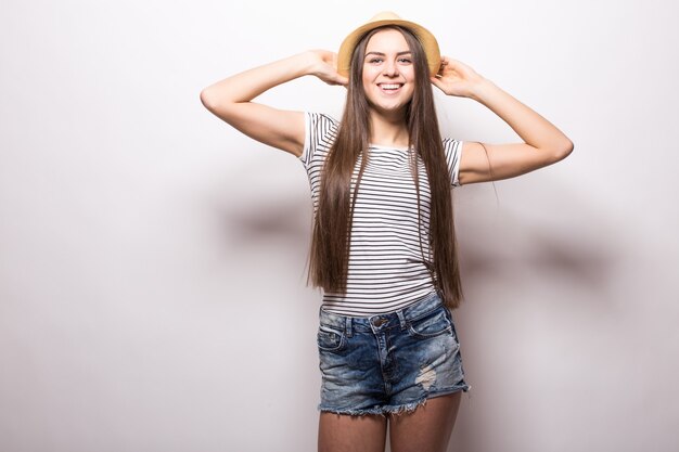 Gorgeous female model keeps hand on straw hat, wears white top with bare shoulders, looks with confident expression, isolated over white wall