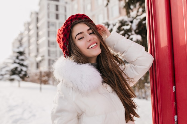 良い気分で雪に覆われた通りでポーズをとってストレートの髪型を持つ豪華な女性モデル。冬の間に楽しんでいるニットの赤い帽子のうれしい淡い女性の屋外の写真