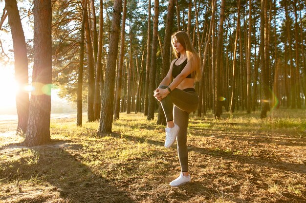 gorgeous female runner doing stretching exercise, workout in the wood in the sun