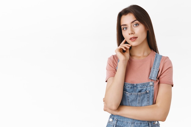 Gorgeous feminine brunette modern girl in dungarees tshirt touching gently cheek and gazing camera with hand crossed over body standing sensual and alluring over white background