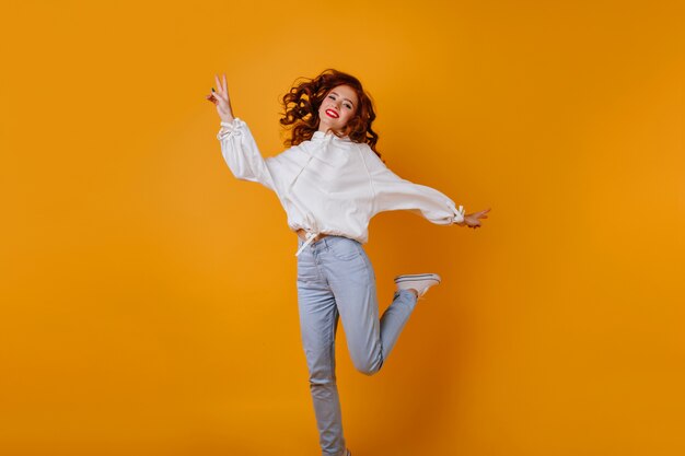 ジャンプするブルージーンズのゴージャスな女の子。オレンジ色の壁で踊る夢のような生姜の女性の屋内写真。