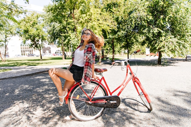 공원에서 여름을 즐기는 체크 무늬 셔츠에 화려한 소녀. 빨간 자전거에 앉아 웃 고 귀여운 여성 모델의 야외 사진.