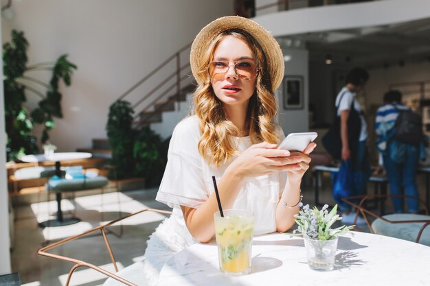 Gorgeous girl with curly hairstyle waiting friend in restaurant with cozy interior and drink icy juice