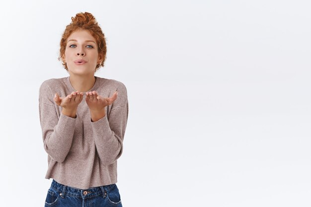 Gorgeous, glamour redhead curly woman combed in messy bun, send kiss at camera, folding lips bend forward and hold hands near mouth to blow mwah, standing tender white wall