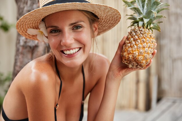 Gorgeous happy female in bikini and summer hat relaxes on spa in tropical hotel, holds pineapple, prepares for party with friends. People, healthy eatting, fruit diet and recreation concept.