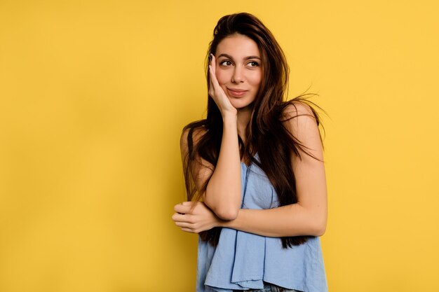 Gorgeous happy woman in blue shirt covering face with hand and dreamily look into the frame on yellow wall.