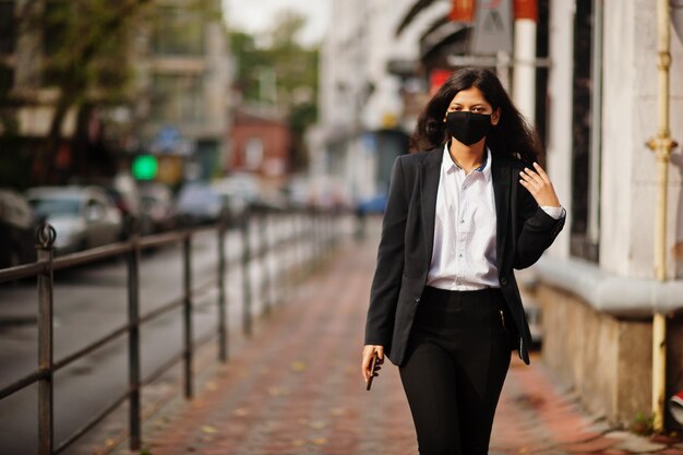 Великолепная индийская женщина в формальной и черной маске для лица позирует на улице во время ковидной пандемии с мобильным телефоном под рукой